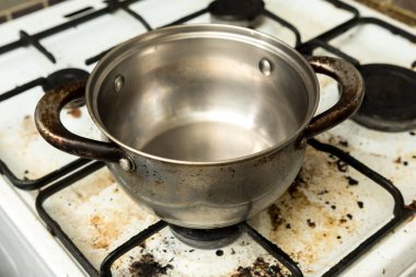 Ocağında yağlı, metal bir tencere olan kirli bir gaz sobası. Görüntü, mutfak hijyen sorunlarını, ev bakımı sorunlarını ve günlük yemek pişirme sorunlarını vurguluyor.