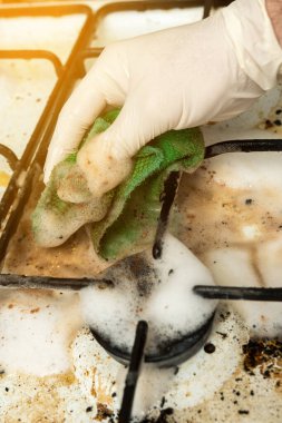 Kirli gaz ocağı köpükle ve yeşil bir bezle eldivenle temizleniyor. Mutfak temizliği rutinindeki çabayı yakalar
