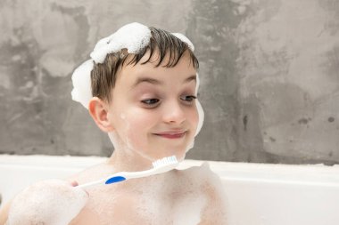 Kafasında ve omuzlarında sabun köpükleriyle banyodan zevk alan küçük bir çocuk, aşağıya bakıyor ve plastik bir diş fırçası tutuyor. Günlük hijyen rutinlerini ve çevre dostu olmayan öğelerin kullanımını vurgular.