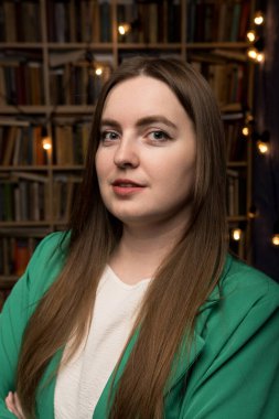 Yeşil ceketli profesyonel bir kadın kitaplarla ve dekoratif ışıklarla dolu bir kitaplığın önünde sıcak ve entelektüel bir atmosfer yaratarak güvenle duruyor..