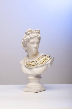 Klasik beyaz mermer büstü, örtülü elbisenin üzerinde karmaşık altın detaylarıyla, mavi arkaplan üzerine kurulmuş. Heykelin yüzü açıkça görülüyor, ayrıntılı özelliklerini sergiliyor..