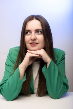 Yeşil ceketli profesyonel bir kadın, elleri çenesinin altına sıkışmış bir şekilde bir masada oturuyor. Kendine güveni ve özgüveni tam. İş, şirket ve profesyonel temalar için mükemmel..