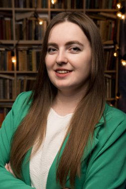 Yeşil ceket giyen profesyonel bir kadın, arkaplanda iyi stoklanmış bir kitaplık ve dekoratif sicim lambalarına karşı çapraz kollarla poz veriyor. Bu da bilimsel ve davetkar bir ortam olduğunu gösteriyor..