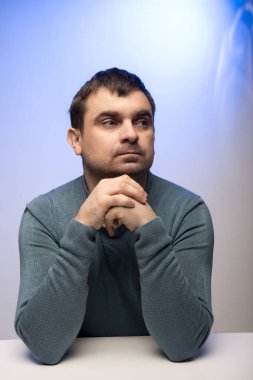 Gök mavisi süveterli bir adam beyaz bir masada elleri sımsıkı bağlanmış ve mavi bir zemine yaslanmış şekilde oturuyor. İş için mükemmel, gündelik, ya da tefekkür teması.