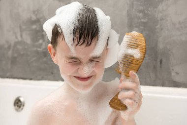 Sabunlu köpüklerle dolu bir küvette saçlarını tahta bir fırçayla tarayan neşeli bir çocuk. Bu neşeli an, banyo sırasında masumiyeti, özgüveni ve eğlenceyi yansıtır.. 