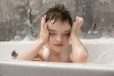 Rahat bir çocuk köpük banyosunun tadını çıkarırken gözleri kapalı ve elleri yüzünde. Huzurlu ve rahatlatıcı bir çocukluk anı, kişisel bakım ve rahatlama. Yumuşak köpük ve sıcak atmosfer.