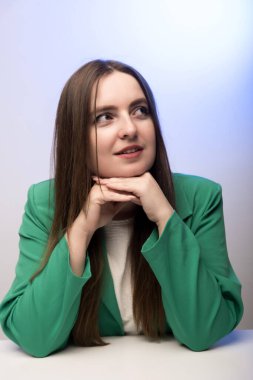 Elleri çenesinde dinlenen yeşil ceketli bir adam, mavi arka planda düşünceli bir poz. Düşünce ve profesyonellik temaları için ideal.