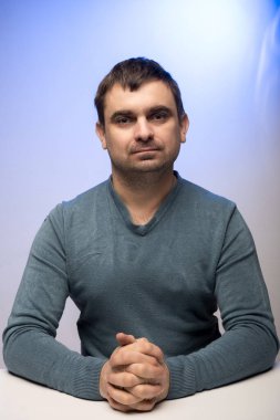 Mavi kazaklı bir adam, elleri bağlı bir masada oturuyor, mavi bir arkaplana karşı. Düşünce, profesyonellik ve resmi ayarlar temaları için uygundur.