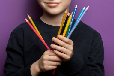 Genç bir öğrenci birden fazla renkli kalem tutuyor, yaratıcılığı, öğrenmeyi ve sanatsal ifadeyi simgeliyor. Parlak mor arkaplanlı yakın çekim.. 