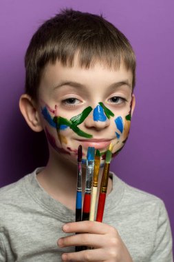 Neşeli çocukların yüzü renkli boyalarla kaplanmış, fırçayla son darbeyi vuruyor. Etkileyici ve sanatsal an çocukluk özgürlüğü, eğlence ve yaratıcı enerjiyi yakalıyor. Mor arkaplan.