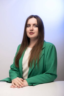 Yeşil ceketli, kendine güvenen bir kadın ellerini mavi bir arkaplanda beyaz bir masaya oturtur. İş, liderlik ya da profesyonel portreler için mükemmeldir..