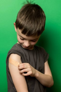 Kolunda bandaj olan bir çocuk, aşağıya bakıyor. Yeşil arka plan. Çocukluk, sağlık, bakım ve iyileşme konuları için ideal.