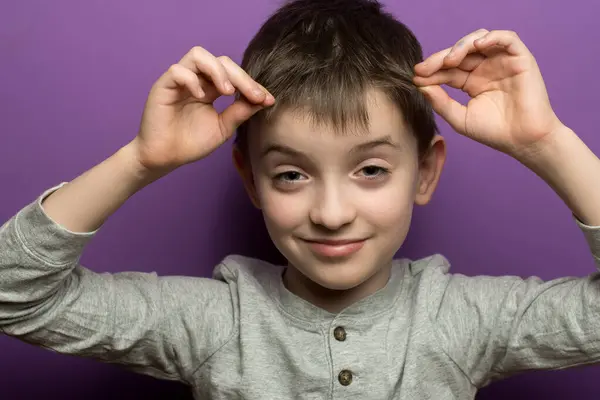 Kısa saçlı çocuk elleriyle tavşan kulağı yapıyor, mor arkaplan. Çocukluk eğlencesi, neşeli jestler, hayal gücü ve masumiyet temaları için uygundur..