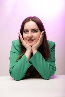 Neşeli bir ifadeyle mutlu bir bayan, yeşil bir ceket giymiş, parlak pembe bir fonda ellerini yaslıyor.