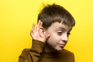 Kahverengi uzun kollu gömlekli meraklı bir çocuk kulağını kaplıyor, sarı arka planda dikkatle dinliyor..