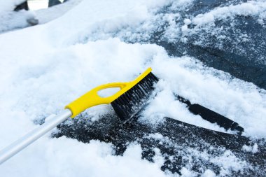 Soğuk hava ve çabayı simgeleyen güneşli bir yerleşim bölgesinde, ön camında sarı kar fırçası olan karlı bir arabanın sakin kış görüntüsü..