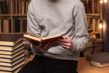 Gri kazaklı bir kişi, kitap rafları ve sıcak aydınlatmalarla çevrili rahat bir kütüphanede bilgi ve sükuneti çağrıştıran kırmızı bir kitap okur..