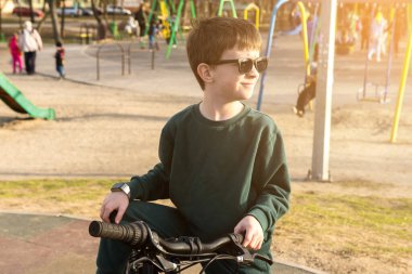 Çocuk, altın saat boyunca parkta bisiklet sürerken oyun yapıları görülebiliyor. Açık hava eğlencesi, aile zamanı ya da aktif çocuklar için harika..