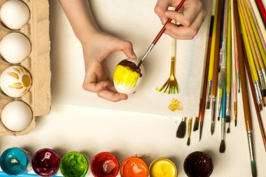 Bir karton içinde renkli boyalar ve yumurtalar bulunan bir Paskalya yumurtasını fırçayla boyayan yakın plan bir el resmi. Şenlikli sanatlar, bahar sanatı veya yaratıcı ifade için uygundur..