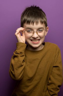 Kahverengi gömlekli ve gözlüklü neşeli bir çocuk mor bir arka plana karşı büyük bir gülümsemeyle gözlerini ayarlar. Eğitim, çocukluk sevinci ya da neşeli anlar için mükemmel..