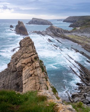 Görüntü, denize doğru uzanan sert bir kaya oluşumunun olduğu engebeli bir kıyı şeridini yakalıyor. Dalgalar kayalara çarpar ve sakin su yüzeyi ile keskin uçurum kenarları arasındaki kontrastı vurgular. Parçalı bulutlu gökyüzü değişmemizi öneriyor.