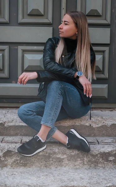 美丽的高加索女孩 金发碧眼 穿着黑色夹克 蓝色牛仔裤站在台阶上 背景是城市风貌 — 图库照片
