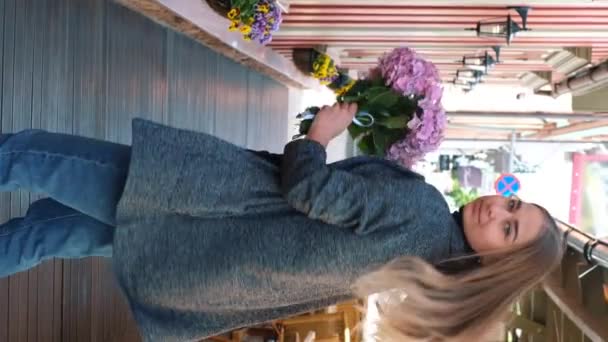ブロンドの髪をした美しい白人の女の子 グレーのコートとピンクのアジサイの花束と背景の街の景色を望むサングラス付きの青いジーンズ 垂直ビデオ — ストック動画