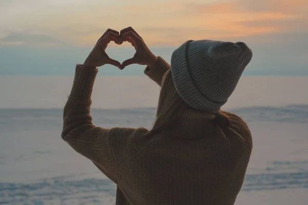A woman making a heart with their hands on the background of frozen sea and sunset.