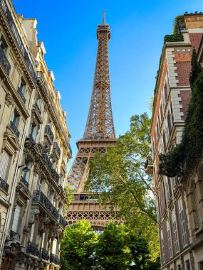 Paris, Fransa 'daki Eyfel Kulesi' nin güzel bir manzarası. Kule ağaçlar ve binalarla çevrili, resim gibi bir manzara yaratıyor. Gökyüzü açık ve mavi.