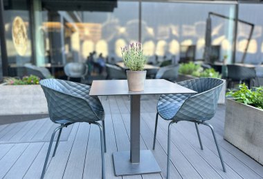 Önünde iki sandalye ve saksı bitkisi olan bir masa. Sandalyeler mavi ve masa ahşaptan yapılmış. Sahne açık havada..