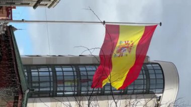 Yüksek bir binanın yanında bir İspanyol bayrağı dalgalanıyor. Bina çok yüksek ve birçok penceresi var. Gökyüzü bulutlu..