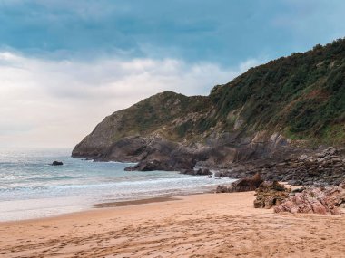 Arka planda bulutlu bir gökyüzü olan kayalık bir sahil. Kumsal sakin ve huzurlu, dalgalar yavaşça kıyıya vuruyor..