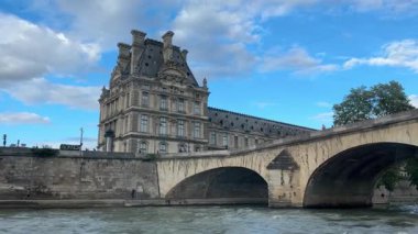 Bir köprü bir nehri kapsar ve arka planda büyük bir bina görülebilir. Gökyüzü açık ve güneş parlıyor. Paris, Fransa.