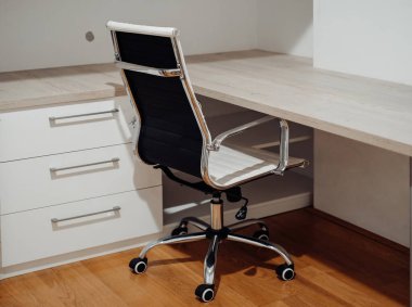 Siyah bir ofis sandalyesi bir masanın önünde oturur. Sandalyenin tekerlekleri var ve tahta bir masanın önünde duruyor.