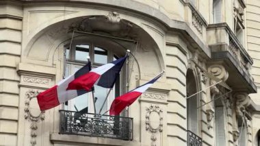 Paris, Fransa 'da tarihi bir taş binanın dekoratif balkonunda sallanan üç Fransız bayrağı. Zarif mimari ayrıntılar kemerli pencereyi çerçeveleyerek klasik Avrupa tarzı ve vatanseverliğini gözler önüne seriyor