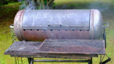 Arka bahçede eski metal barbekü ızgarası. Duman yükseliyor. Yeni veya devam eden aşçılığı gösteriyor. Altında ızgara fırçası olan paslı ve yıpranmış bir yüzey. Açık yeşil çimenler ve bulanık ağaçlar