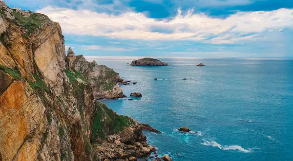 Turkuaz denize bakan kıyı uçurumları ve canlı bulutlu bir gökyüzünün altında kayalık adalar. Seyahat, doğa ve macera temaları için ideal deniz manzarası.