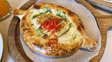 Kızarmış peynir, susam tohumu, şifalı otlar ve dilimlenmiş kırmızı biberle kaplı Gürcü khachapuri. Ahşap bir tahtada servis edilen geleneksel tekne biçimli ekmek, yemek, seyahat ve kültür temaları için mükemmel.