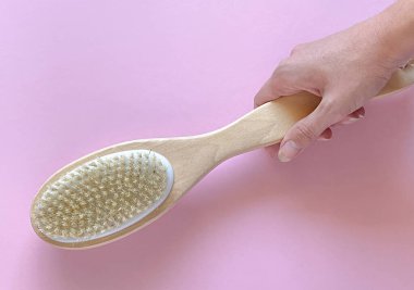 Yumuşak pembe bir arka plana yerleştirilmiş, tahta saplı doğal kıllı bir vücut fırçasını tutan elin yakın çekimi. Bu minimal ve temiz kompozisyon güzellik, cilt bakımı, sağlık, kaplıca veya