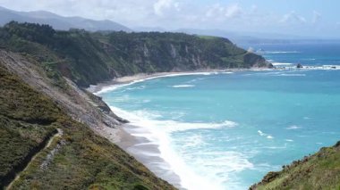 İspanya, Asturias 'ın nefes kesici kıyı manzarası. Turkuaz okyanus dalgalarına ve sakin kumlu plajlara doğru dramatik uçurumlar iniyor. Parlak gökyüzünün altındaki yemyeşil tepeler ve uzak dağlar