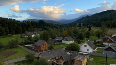 Ukrayna 'daki manzaralı dağ köyü akşam saatlerinde yemyeşil ormanlar ve yuvarlanan tepelerle çevrilidir. Geleneksel ahşap ve taş evler, üzerlerindeki dramatik bulutlu gökyüzü ile yeşil manzarayı noktalıyor.