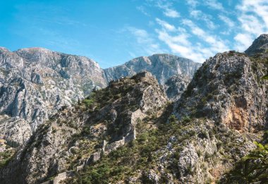 Antik taş kale duvarının panoramik görüntüsü berrak mavi gökyüzünün altındaki engebeli kayalık dağların arasından süzülüyor. Dramatik manzara ve tarihi mimari zamansızlık hissi yaratıyor.