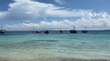 Zanzibar 'da turkuaz tropikal sularda yüzen geleneksel ahşap teknelerin manzarası dramatik beyaz bulutlarla dolu açık mavi bir gökyüzünün altında. Huzurlu sahil sahnesi güzelliği ve sükuneti yakalar.