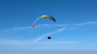 Renkli gökkuşağı gölgesiyle parlak mavi gökyüzüne karşı zarif bir şekilde süzülen bir paraglider. Görüntü, açık bir günde yamaç paraşütünün özgürlüğünü ve heyecanını yakalıyor. Macera ve uçuşu simgeliyor..