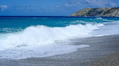 Güçlü okyanus dalgaları, parlak mavi bir gökyüzünün altındaki çakıl taşlı bir sahile çarpıyor. Arka planda engebeli bir uçurum ve uzak bir deniz feneri var. Asturias, İspanya. Turkuaz ve koyu mavi renkler