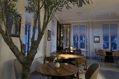 Modaya uygun, modern bir kafe ya da restoranın minimalist estetik bir iç çekimi. Yuvarlak ahşap masalar ince ışıklandırma ve küçük vazolarla döşenmiş, rahat ve misafirperver bir ortam yaratıyor.