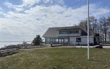 İki katlı, büyük pencereli, muhtemelen bir restoran ya da sakin sulara bakan bir konut binalı, huzurlu bir göl manzarası. Sahne parçalı bulutlu bir gökyüzünün altında geçiyor.