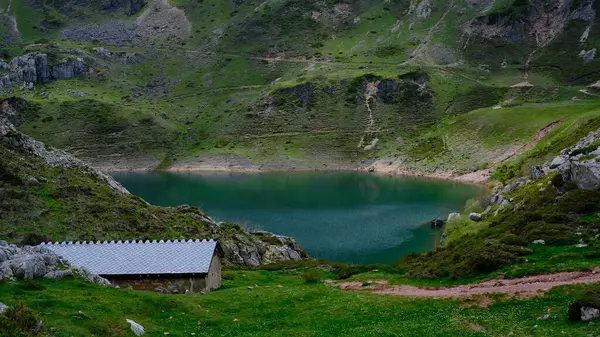 İspanya 'nın Asturias bölgesindeki Somiedo Doğal Parkı' nda yeşil bir vadi içinde sakin, zümrüt yeşili bir dağ gölü bulunur. Gri çatılı geleneksel bir taş kulübe ön plandaki çimenli yamaçlarda oturur.