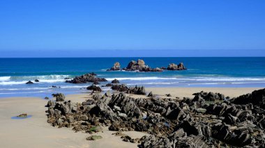 İspanya, Asturias 'ta geniş açılı, parlak bir kıyı manzarası, açık, mavi bir gökyüzünün turkuaz okyanus ufkuyla buluşması. Ön planda engebeli, pürüzlü, karanlık bir kaya var.