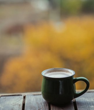 A green enamel mug filled with a warm beverage sits on a rustic wooden surface, surrounded by soft autumn hues. Cozy Autumn Retreat. clipart